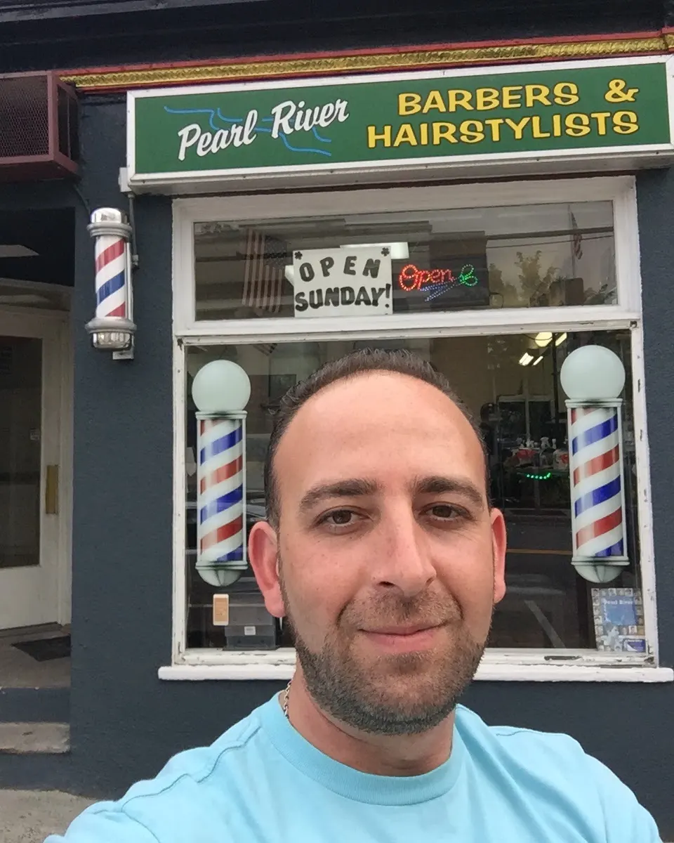 Alex outside Pearl River Barber Shop on Central Ave, barber poles and the Open Sunday sign behind him