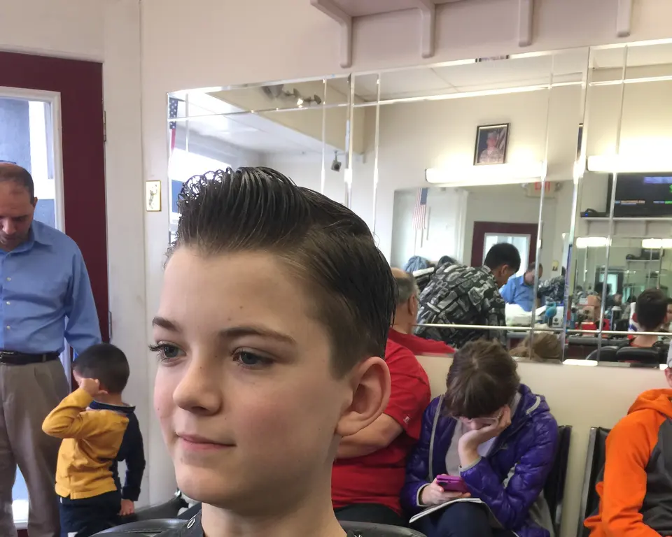 A boy with a fresh slicked-back cut in the chair at Pearl River Barber Shop, families waiting behind